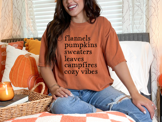 Woman wearing a 'flannels pumpkins sweaters leaves campfires cozy vibes' shirt in a cozy living room.