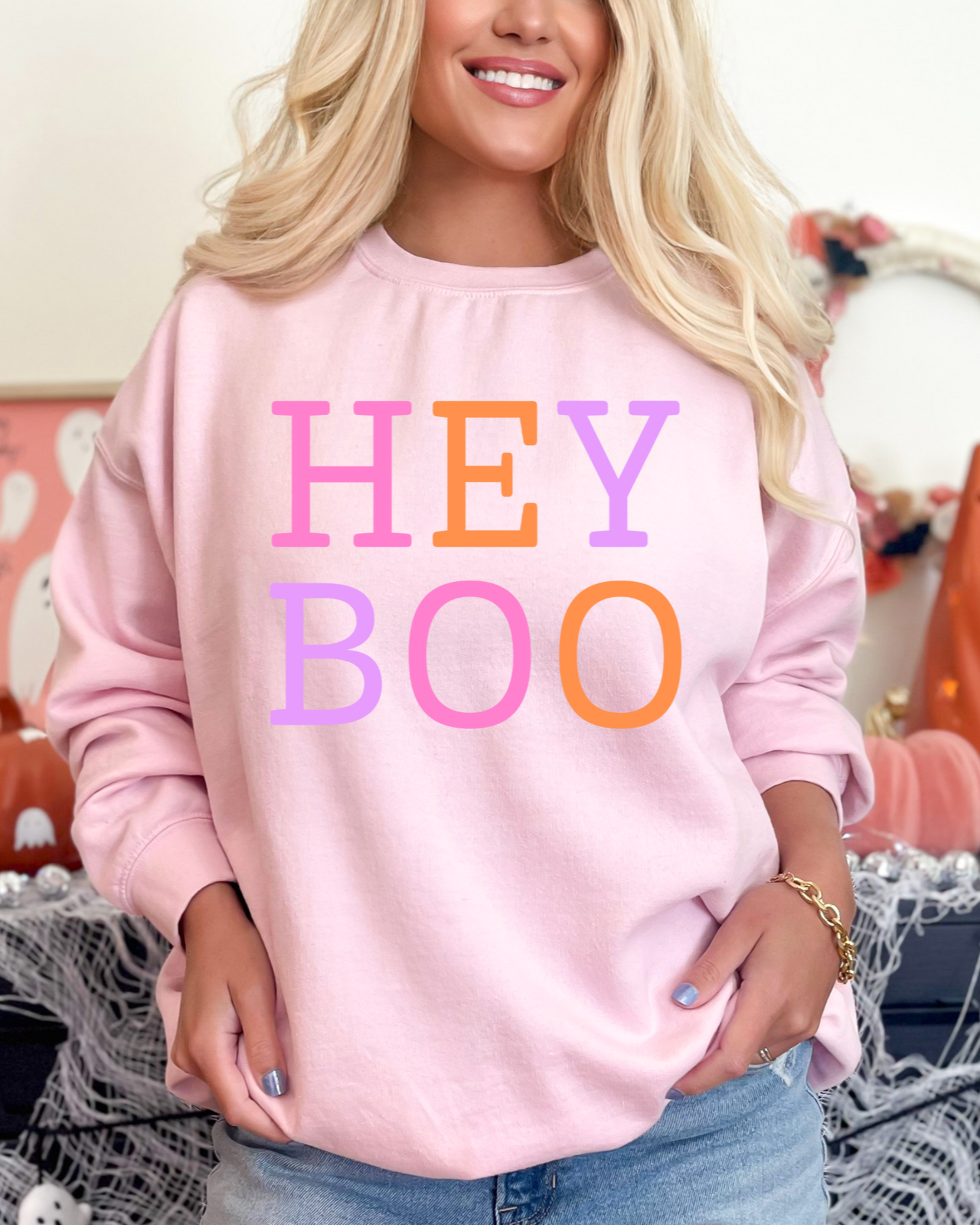 Person wearing a pink sweatshirt with 'HEY BOO' text in colorful letters, standing in a Halloween-themed setting.