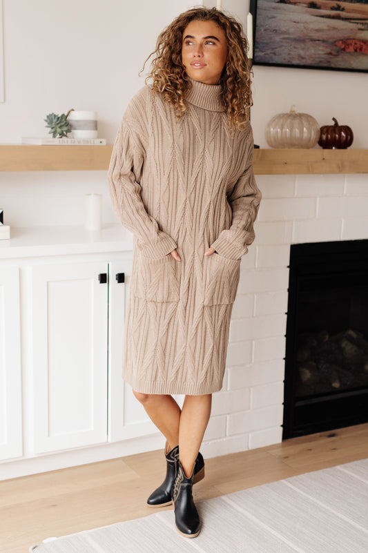 Woman wearing a beige cable knit sweater dress standing in a room with a fireplace.