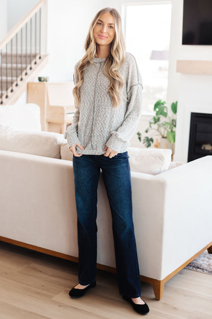 Woman standing in a living room wearing a gray sweater and blue jeans.