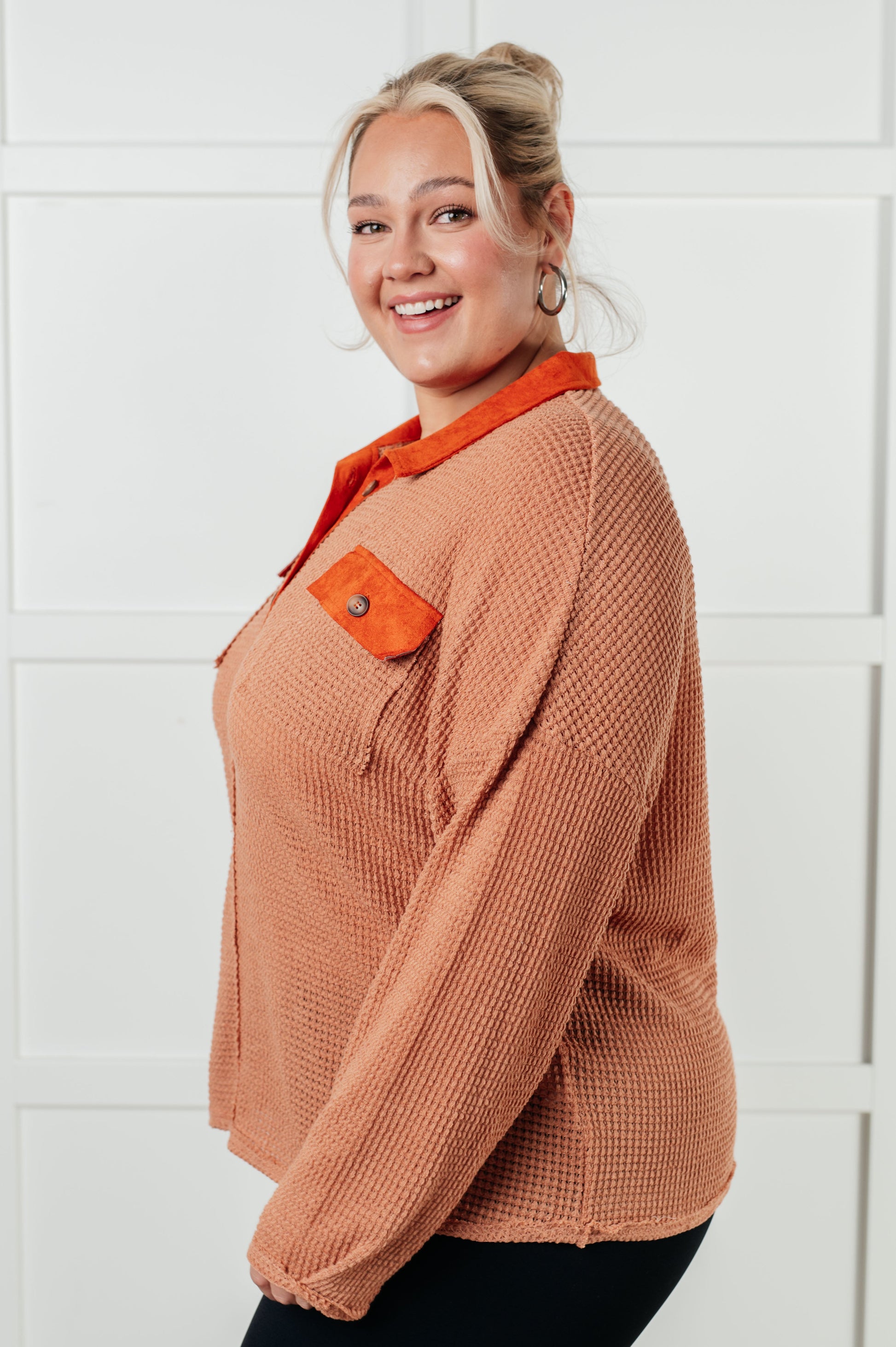 Woman wearing a brown sweater with orange accents against a white tiled wall.