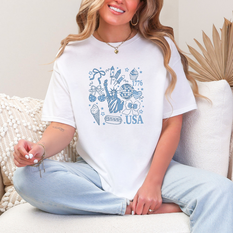 A person sitting on a cushioned chair wearing a white graphic tee with a multicolored geometric pattern and the text 'USA' prominently displayed on the front.