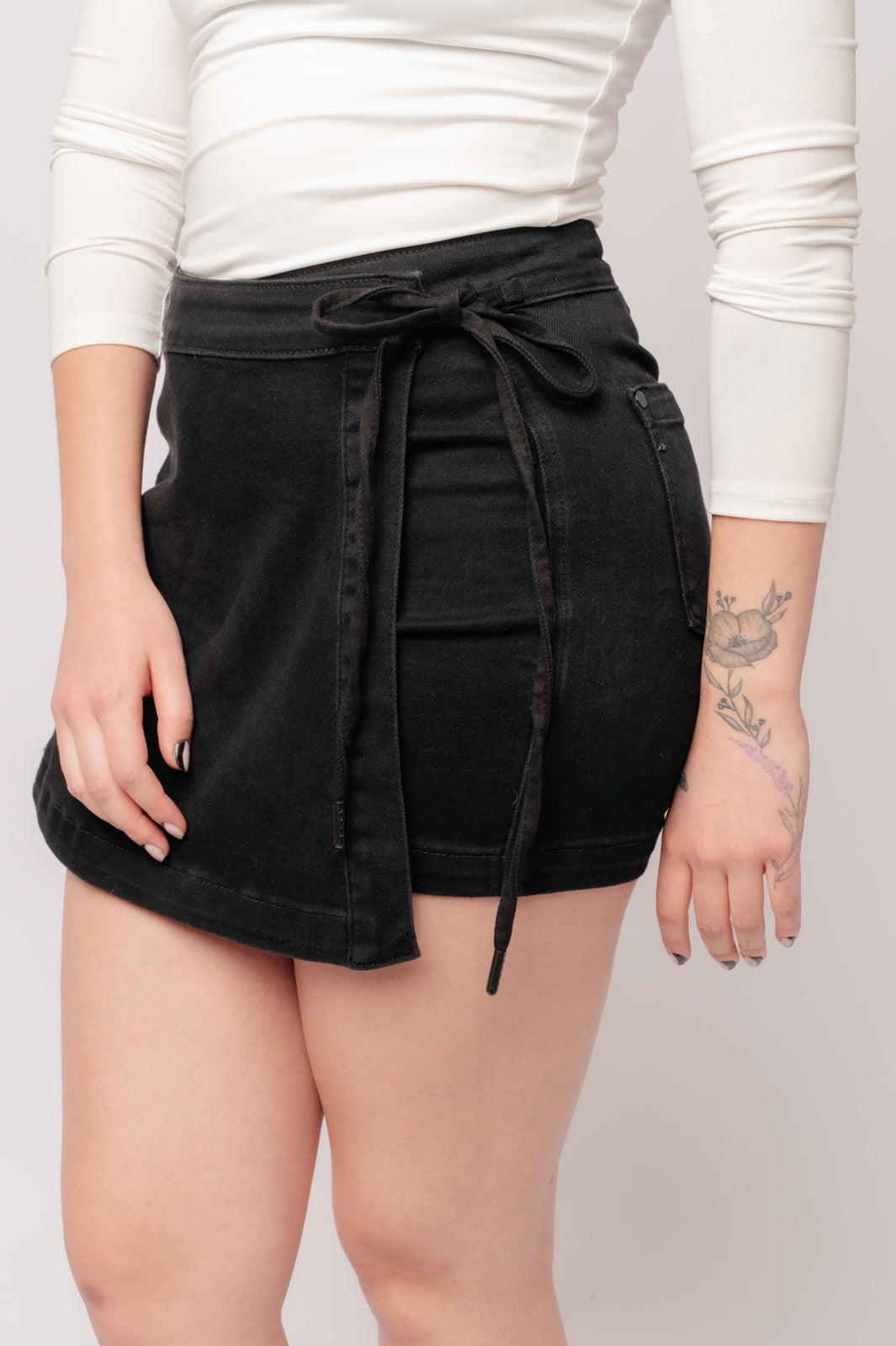A person wearing a high rise side tie denim skort in black color with a self-tie side closure.