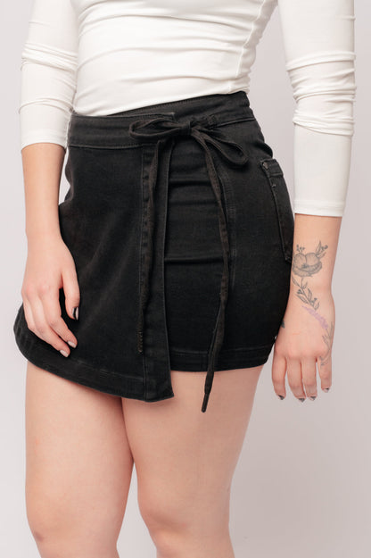 A person wearing a high rise side tie denim skort in black color with a self-tie side closure.