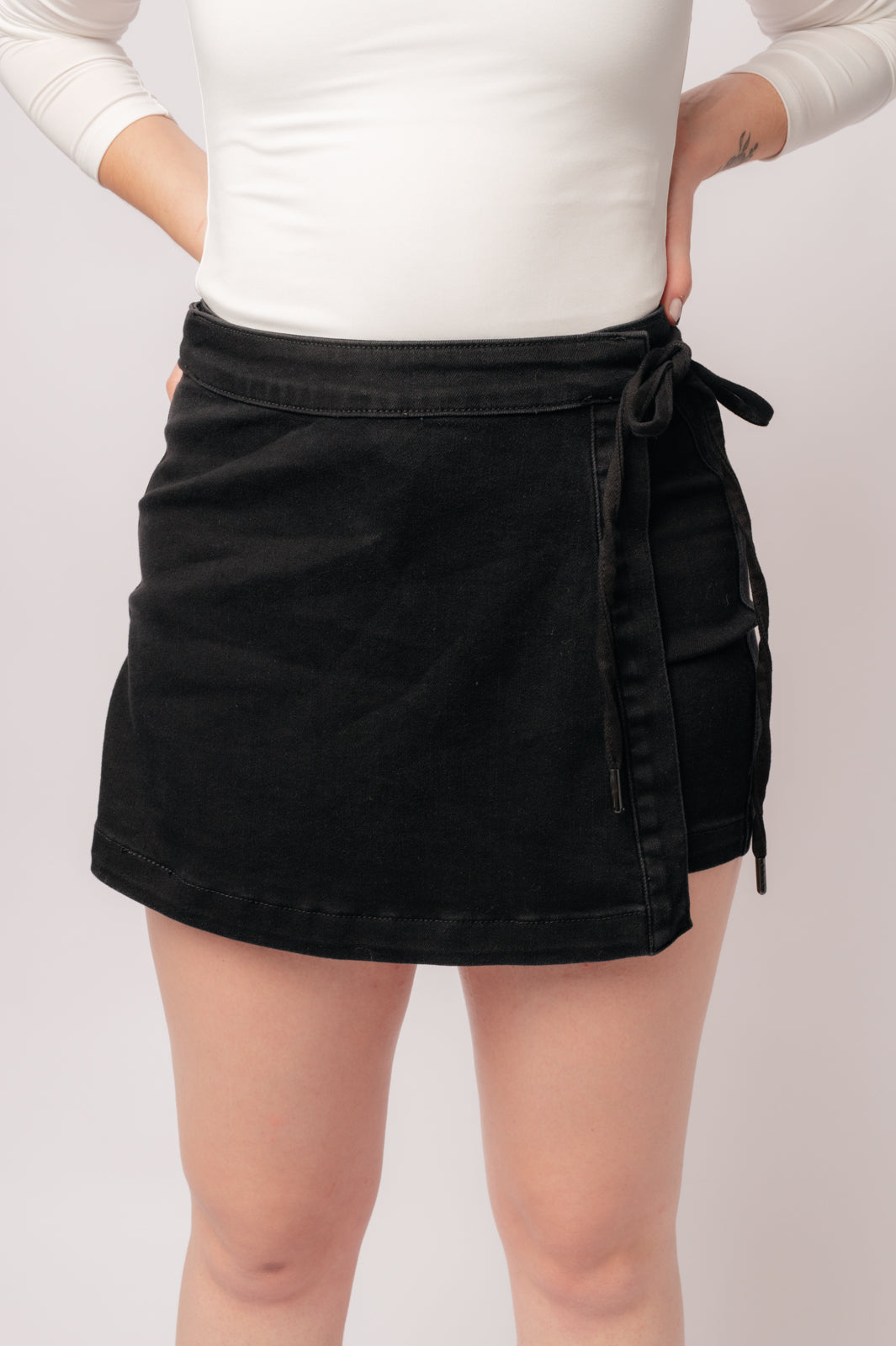 A person wearing a high rise side tie denim skort in black color with a self-tie side closure.
