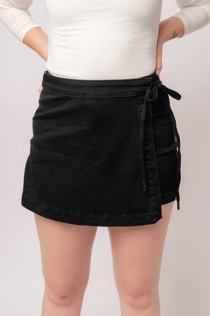 A person wearing a high rise side tie denim skort in black color with a self-tie side closure.