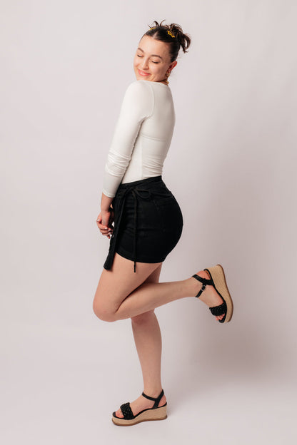 A person wearing a high rise side tie denim skort in black color with a self-tie side closure.