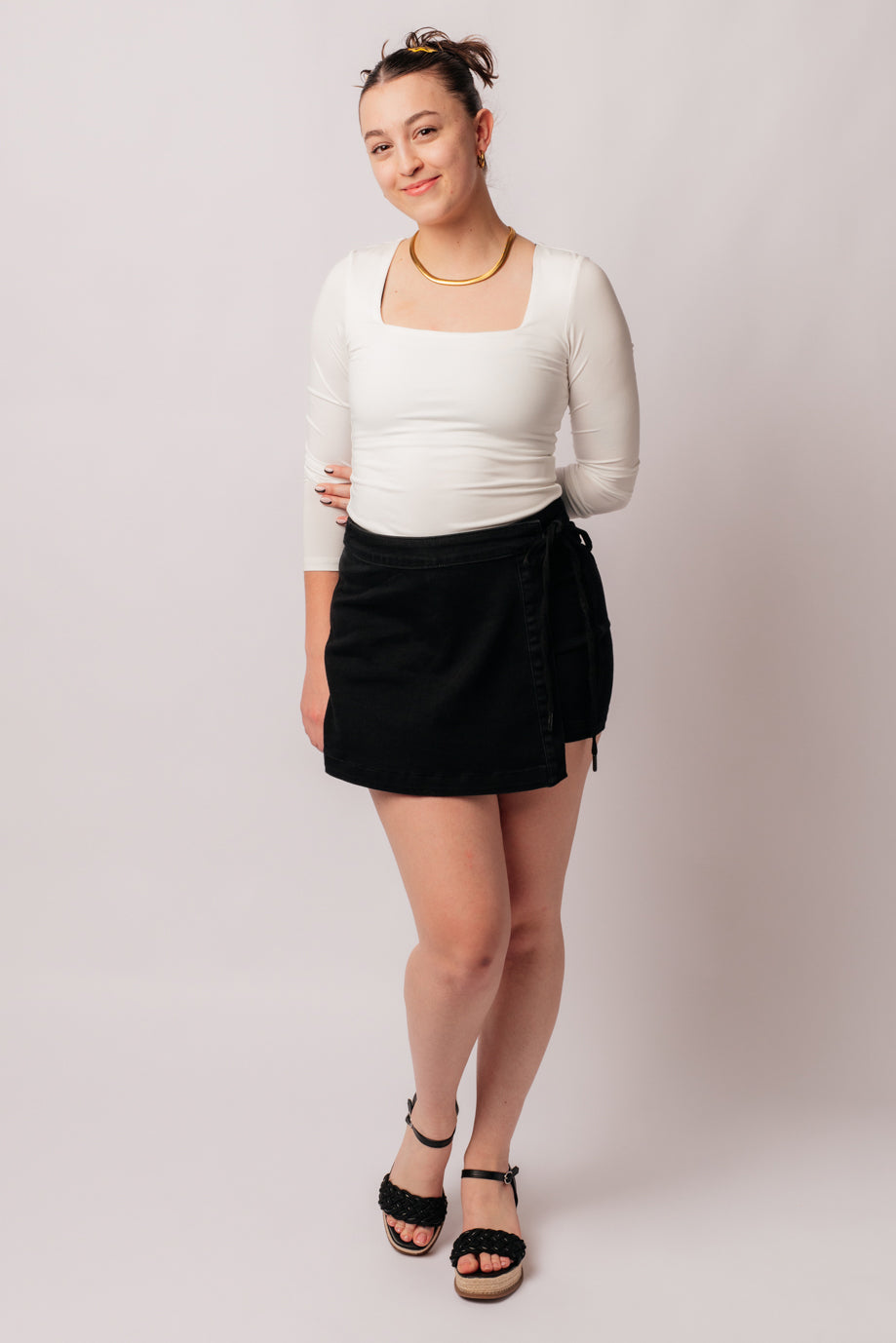 A person wearing a high rise side tie denim skort in black color with a self-tie side closure.