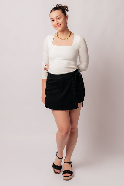 A person wearing a high rise side tie denim skort in black color with a self-tie side closure.
