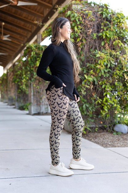 Woman wearing a black long-sleeve top and patterned leggings standing on a sidewalk with greenery in the background.