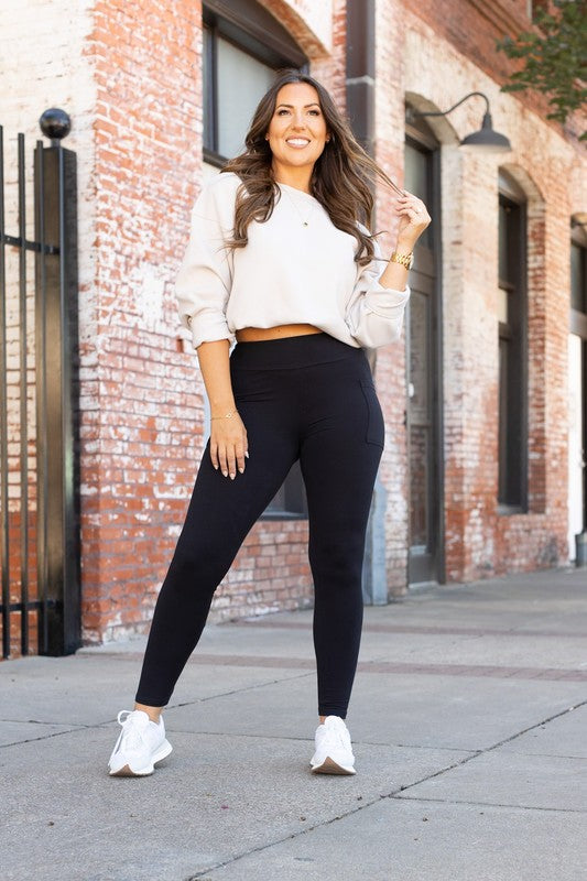 Woman wearing a white blouse and black leggings standing on a sidewalk with a brick building in the background.