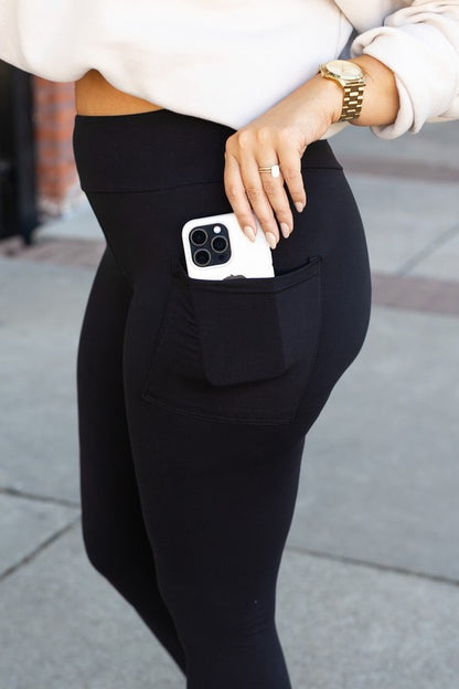 Person wearing black leggings with a pocket holding a phone, standing on a sidewalk.