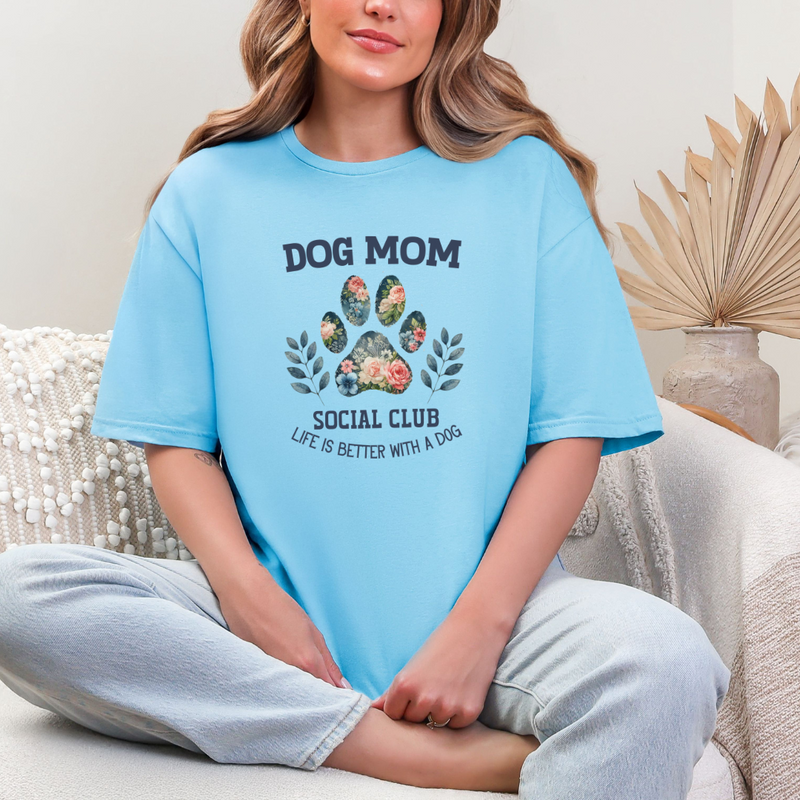 A woman sitting on a couch, wearing a blue graphic tee with 'DOG MOM SOCIAL CLUB' printed on the front.
