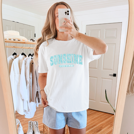 A person wearing a white graphic tee with 'Sunshine State of Mind' printed in blue, short blue shorts, and white shoes, standing in a room with clothes in the background.