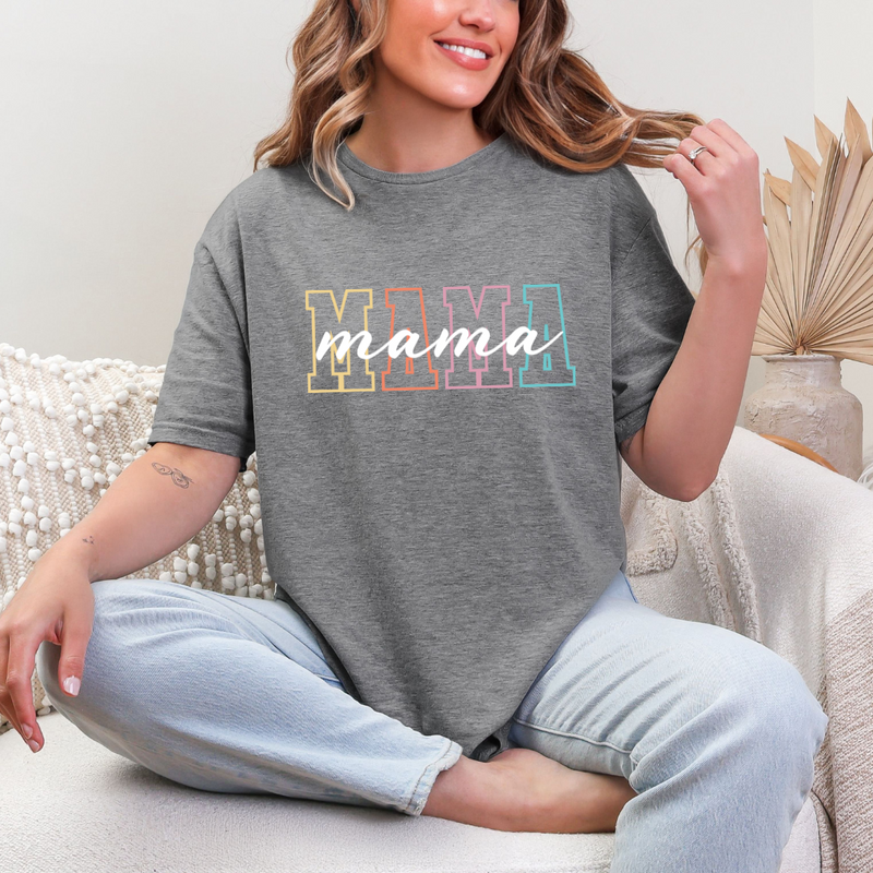 A woman sitting and smiling while wearing a gray t-shirt with the word 'mama' printed in multicolored letters.