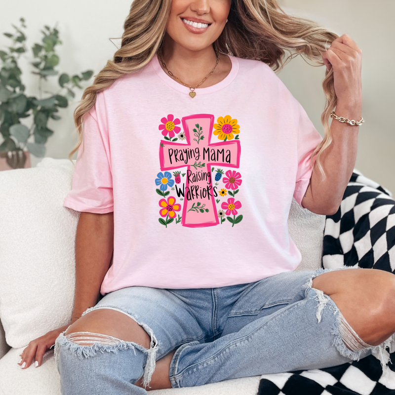 A woman sitting on a couch wearing a pink graphic tee with a floral design and text that reads 'Praying Mama Raising Warriors'.