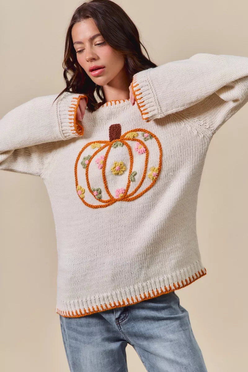 Woman wearing a cream sweater with embroidered pumpkins and flowers on a beige background