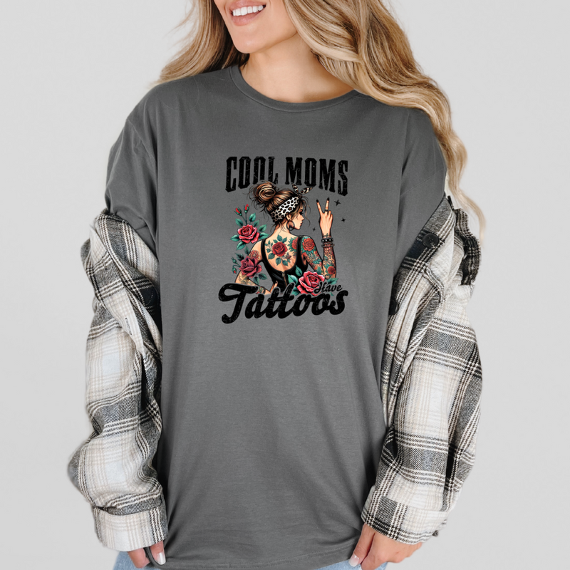 A woman wearing a gray graphic tee with the text 'COOL MOMS HAVE TATTOOS' and a floral design. The tee is crew neck with short sleeves, and she has a plaid shirt tied around her waist.