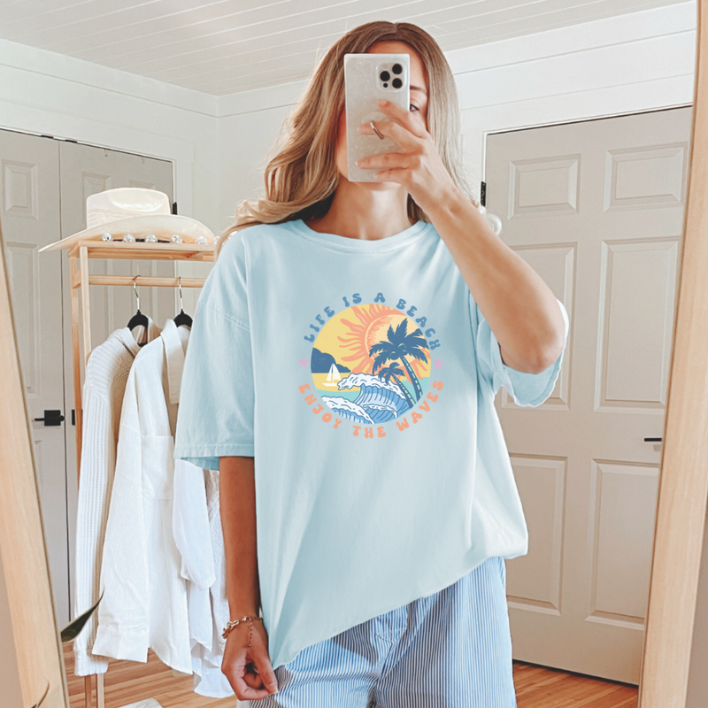 A person taking a mirror selfie wearing a blue graphic tee with a sun and beach-related design.