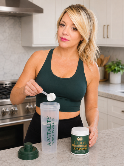 A woman in a dark green sports bra and black leggings stands in a kitchen, holding a spoon over a clear shaker bottle with a green cap. She is next to a green and white container labeled A-VITALITY Core.