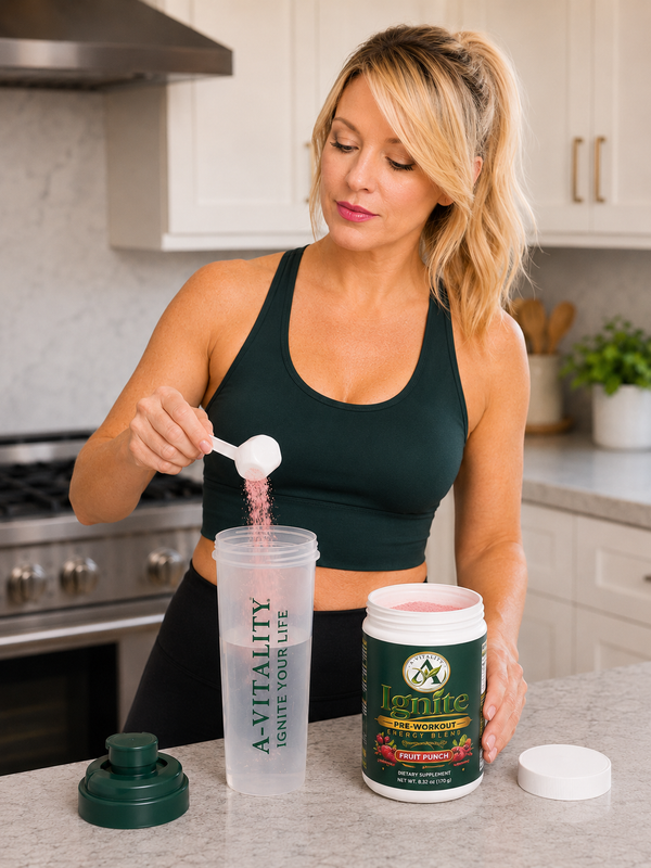 A woman in a dark green sports bra and black leggings stands in a bright kitchen, adding pink powder from a white scoop into a clear shaker bottle. On the counter in front of her are a green-labeled tub of Ignite protein powder, its white lid, and the bottle's green cap.