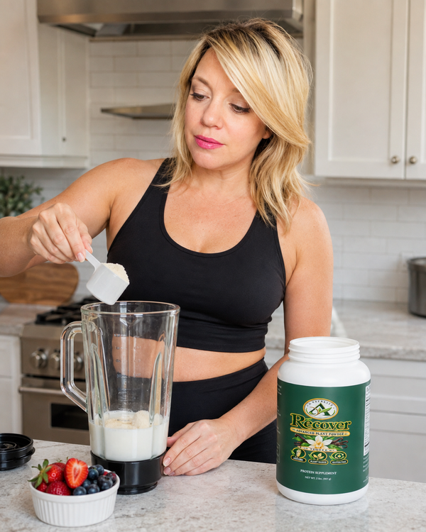 A woman in a black sports bra and leggings mixes powder into a blender in a bright kitchen, with a tub of Recover Protein powder on the counter next to a small bowl of fresh strawberries and blueberries.