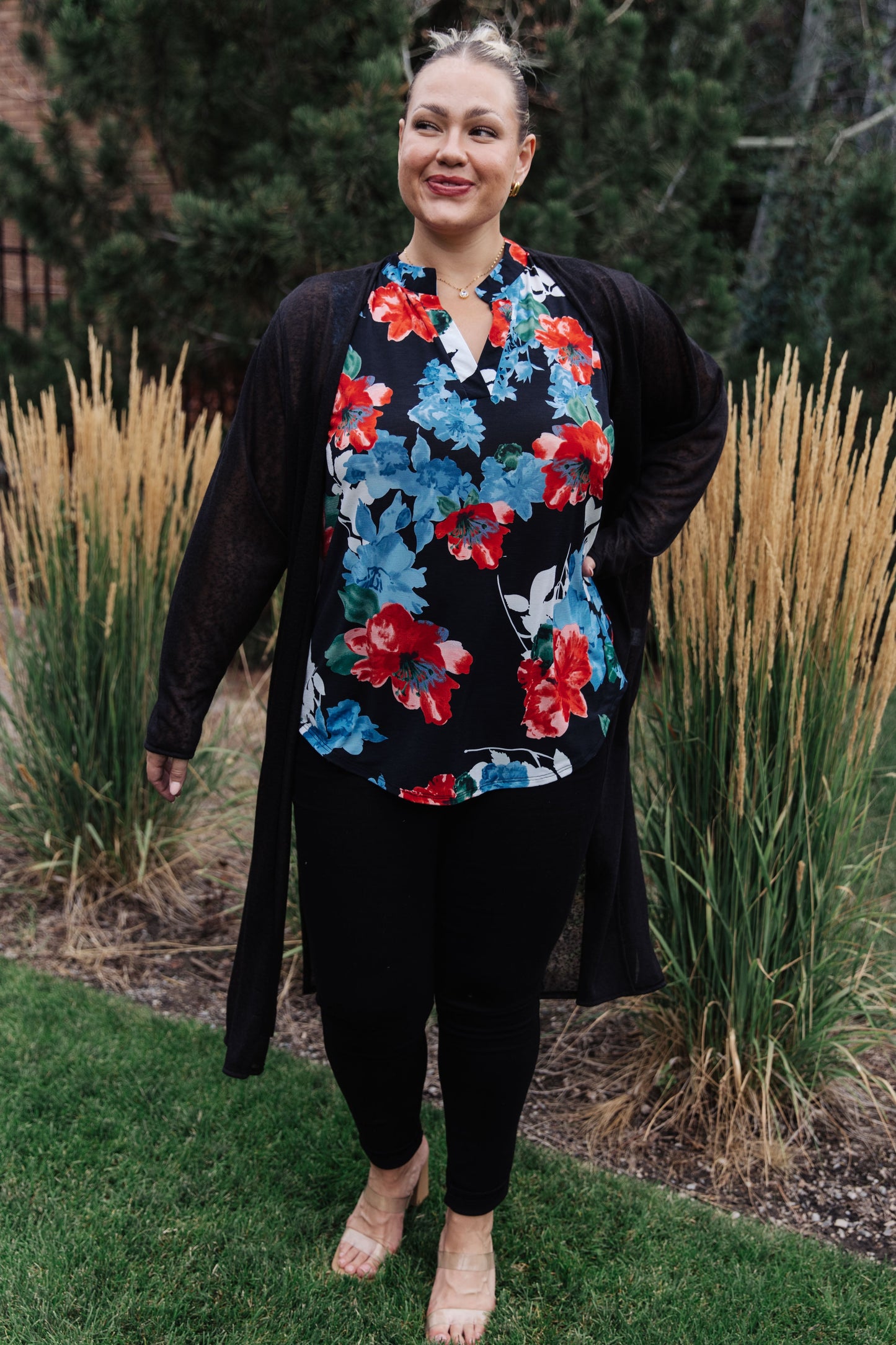 Woman wearing a floral top and black cardigan standing outdoors with tall grasses and trees in the background.