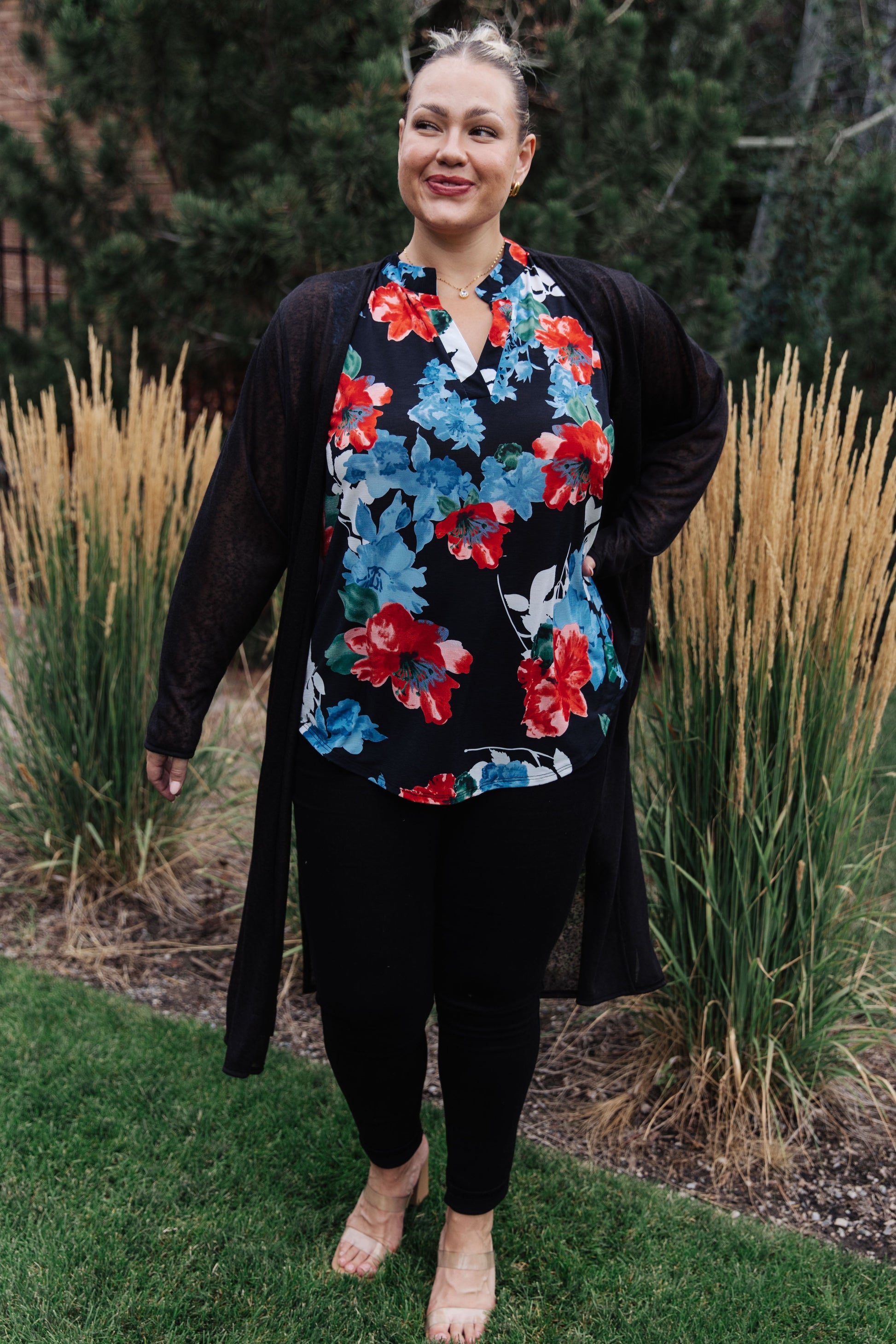 Woman wearing a floral top and black cardigan standing outdoors with tall grasses and trees in the background.