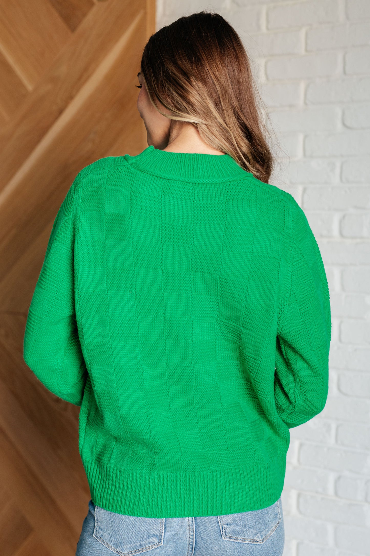 Person wearing a green sweater standing against a white brick wall.