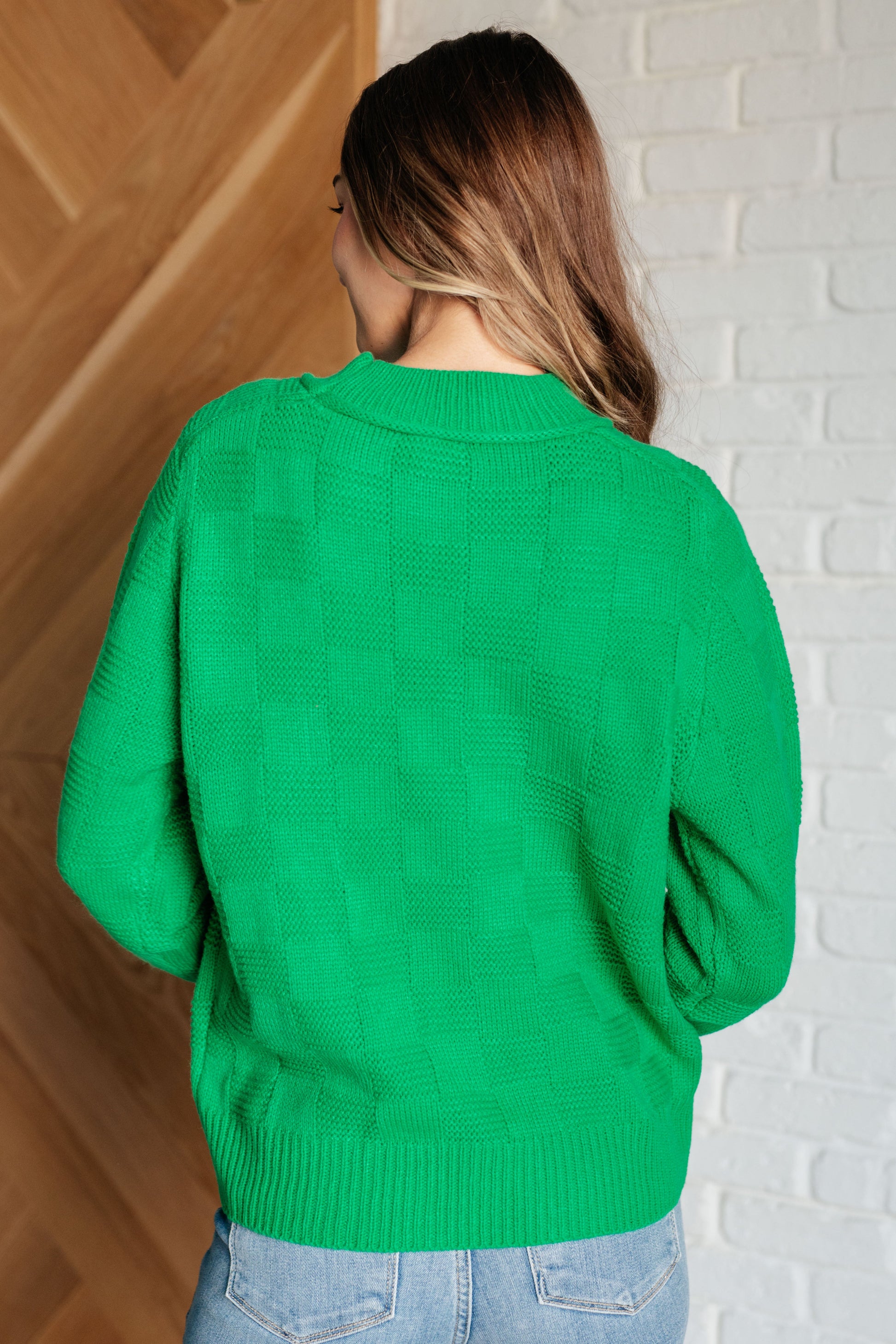 Person wearing a green sweater standing against a white brick wall.