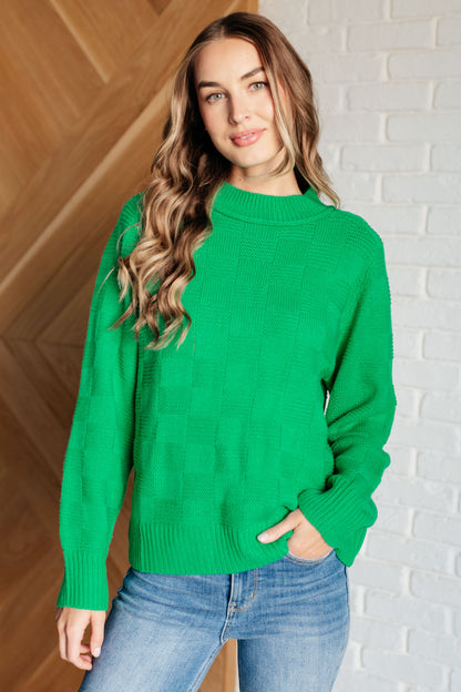 Woman wearing a green sweater and blue jeans against a white brick wall.