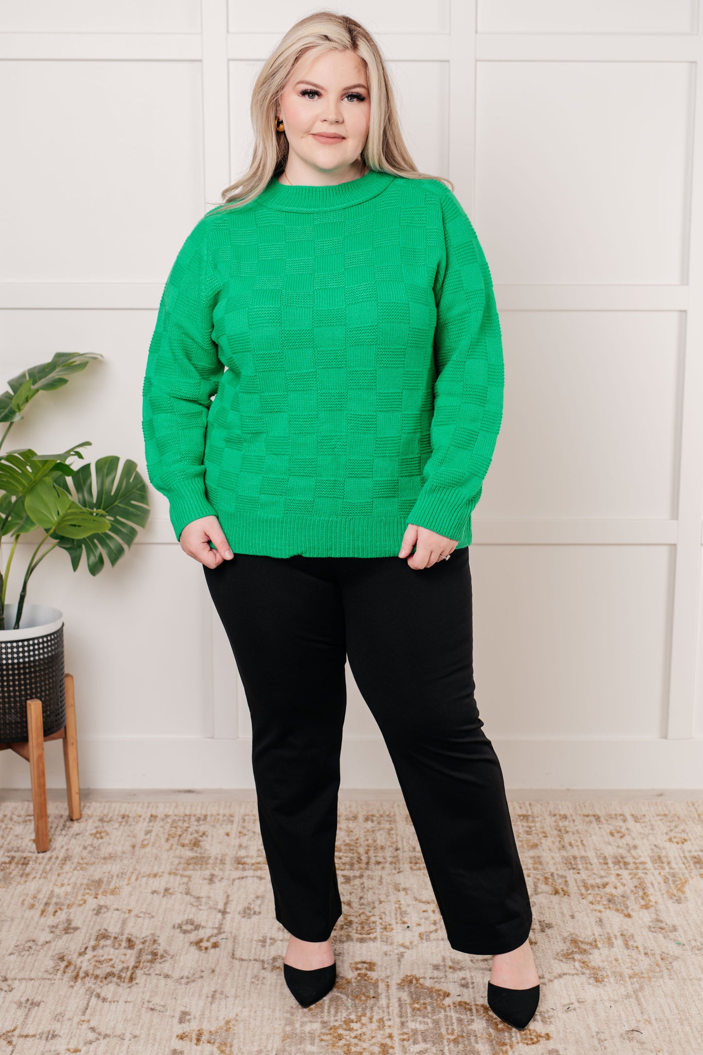 Woman wearing a green sweater and black pants standing in a room with a plant and rug.