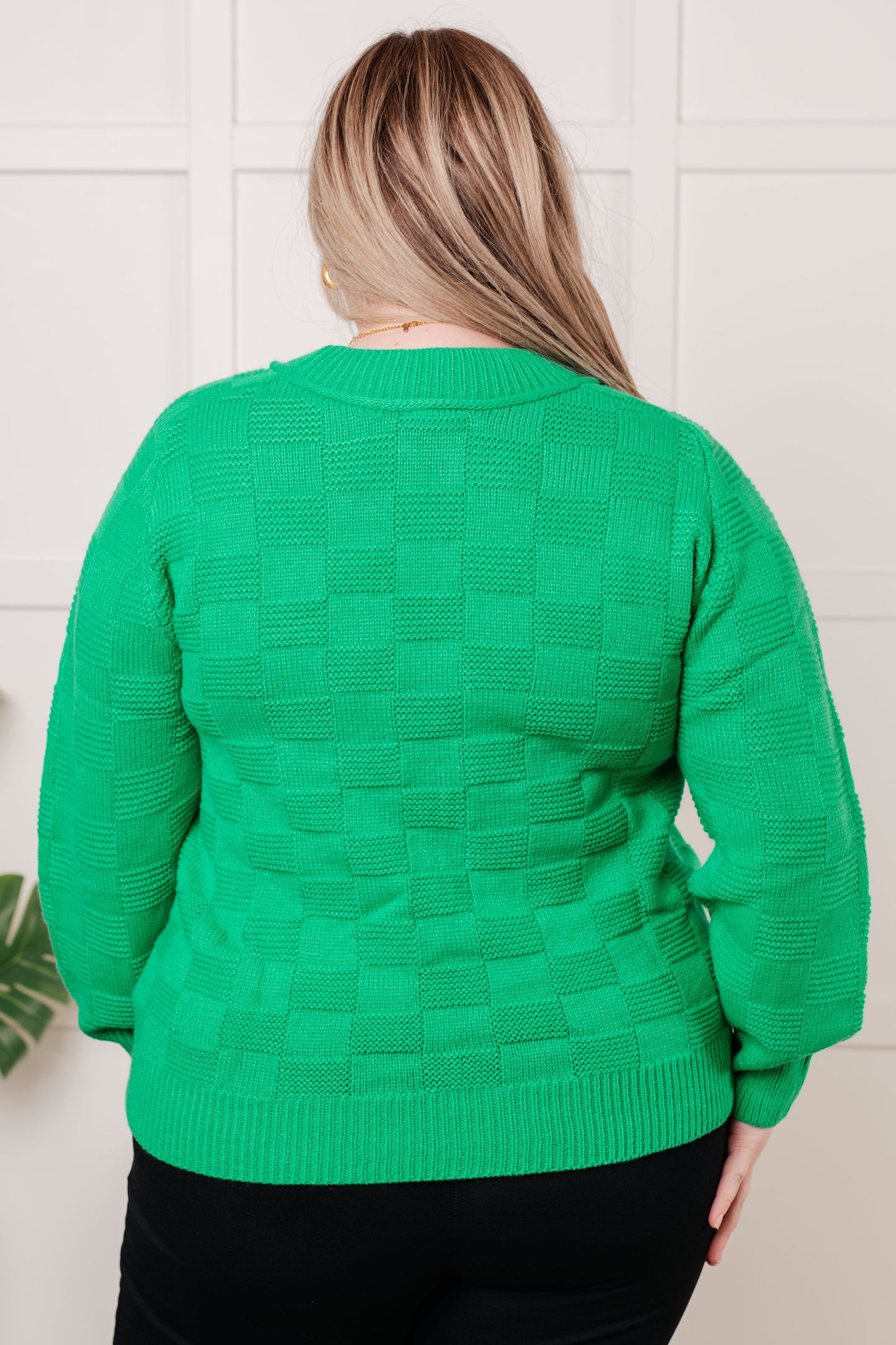 Person wearing a green sweater with a textured pattern against a white tiled wall.