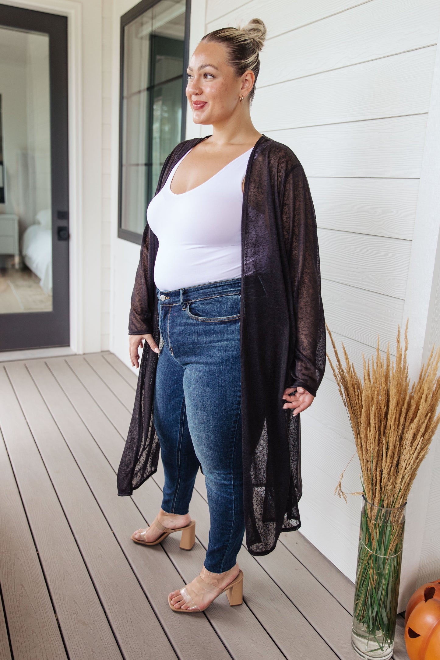 Woman standing on a wooden deck wearing a black sheer cardigan, white top, and blue jeans.