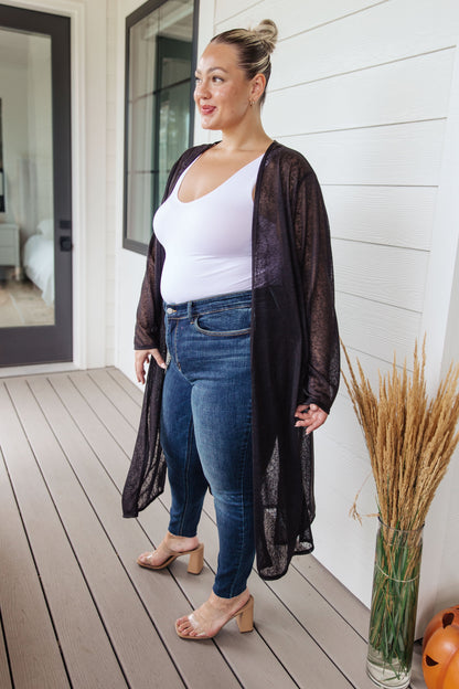 Woman standing on a wooden deck wearing a black sheer cardigan, white top, and blue jeans.