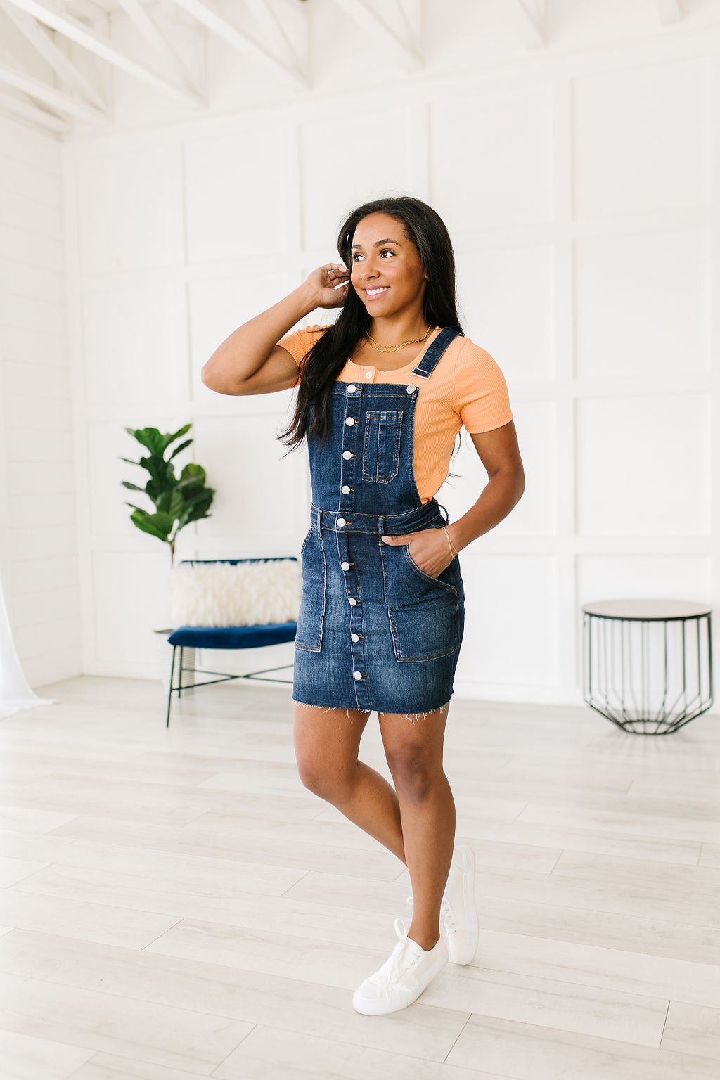 Woman wearing a denim pinafore dress in a bright room.