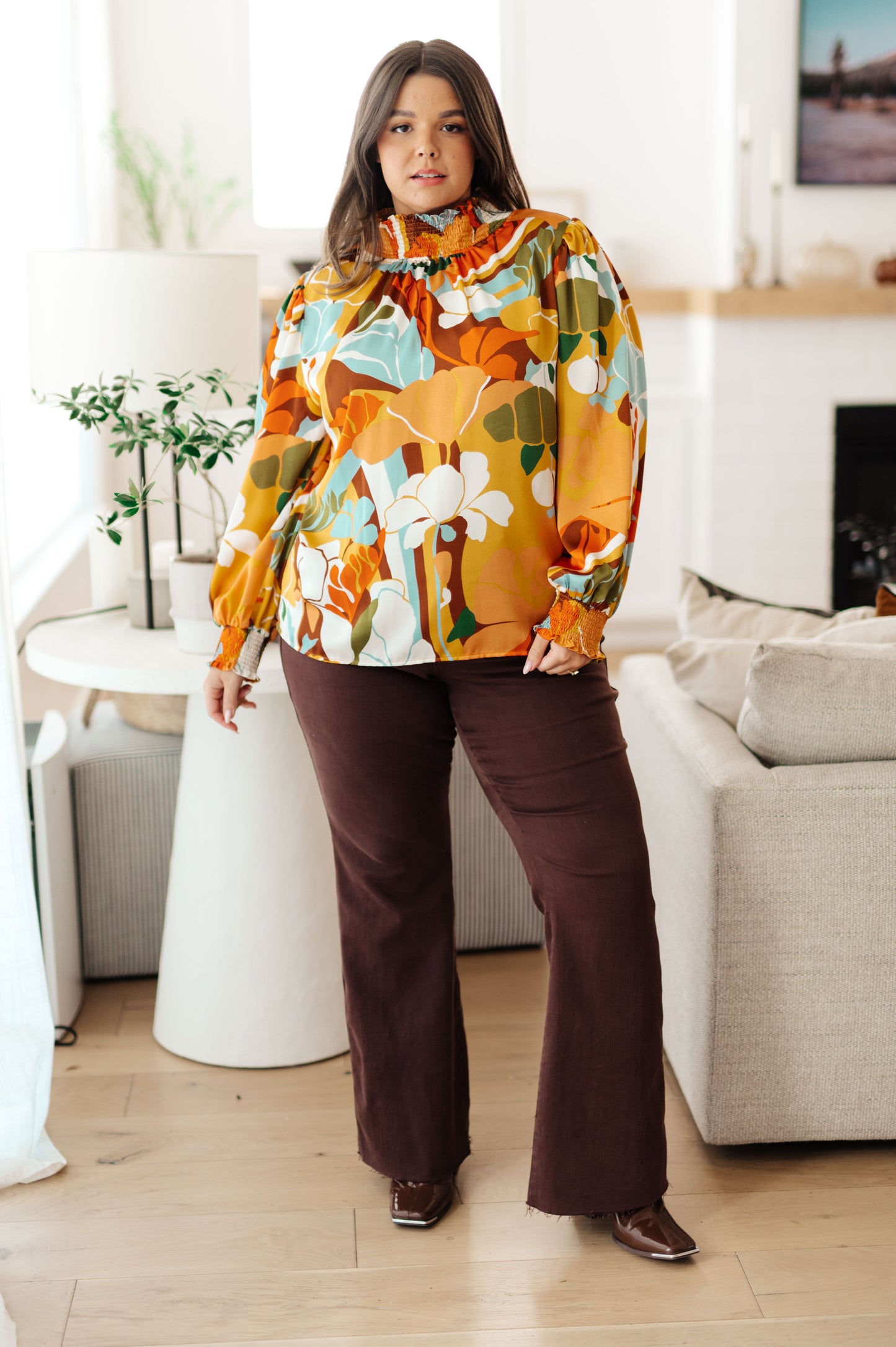 Woman wearing a colorful floral blouse and brown pants in a living room.