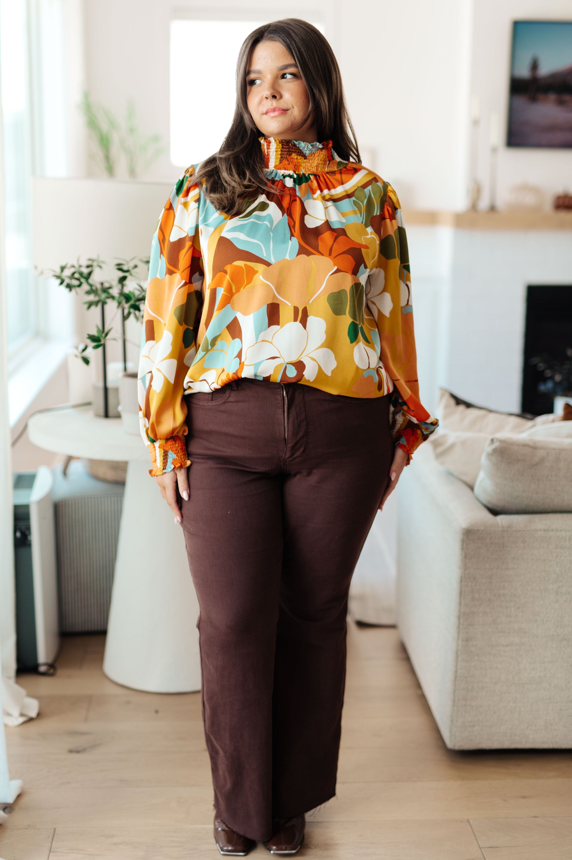 Woman wearing a colorful floral blouse and brown pants in a living room.