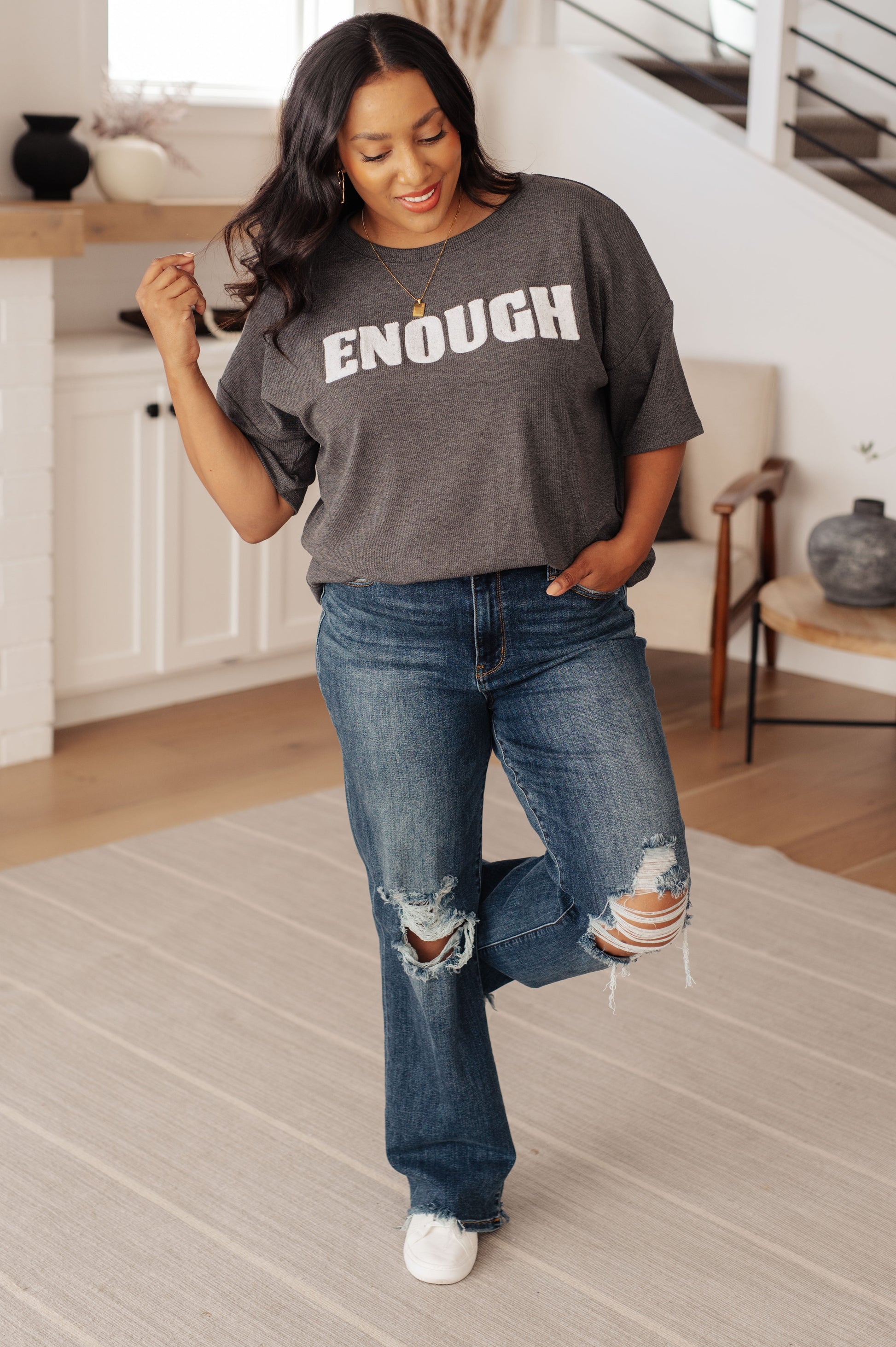 Woman wearing a gray t-shirt with 'ENOUGH' text and blue jeans in a home setting.