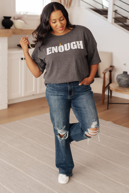 Woman wearing a gray t-shirt with 'ENOUGH' text and blue jeans in a home setting.