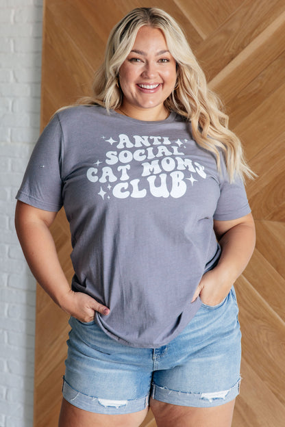 Woman wearing a gray t-shirt with 'Anti Social Cat Mom Club' text, standing against a wooden wall.