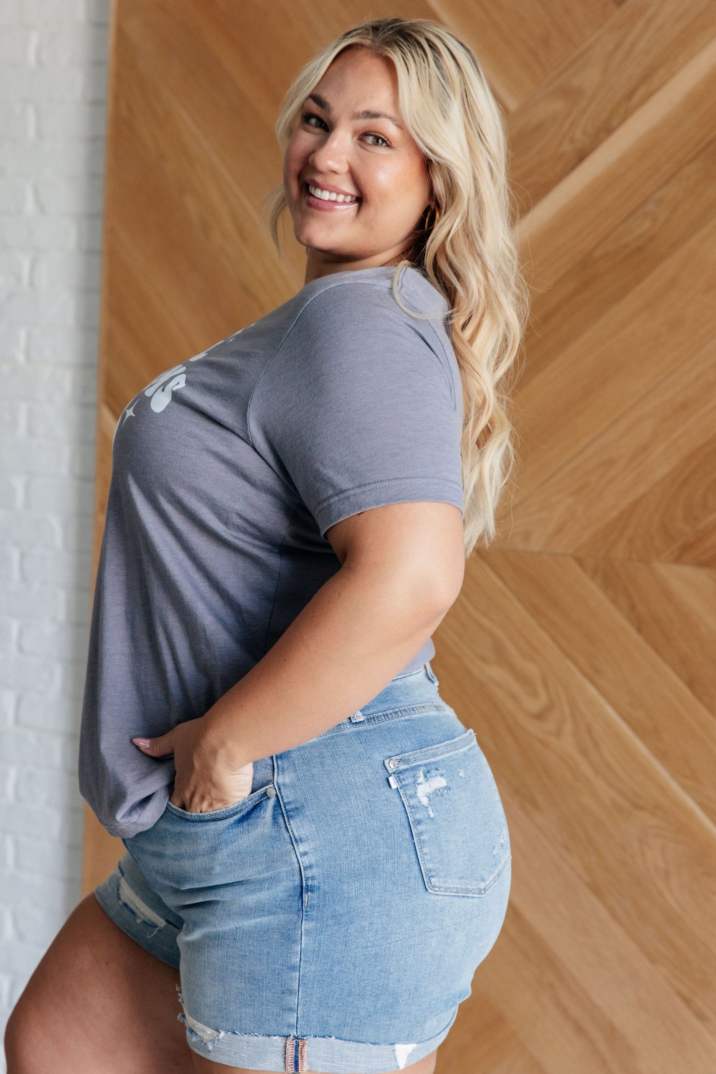 Woman wearing a gray t-shirt and denim shorts standing against a wooden wall.