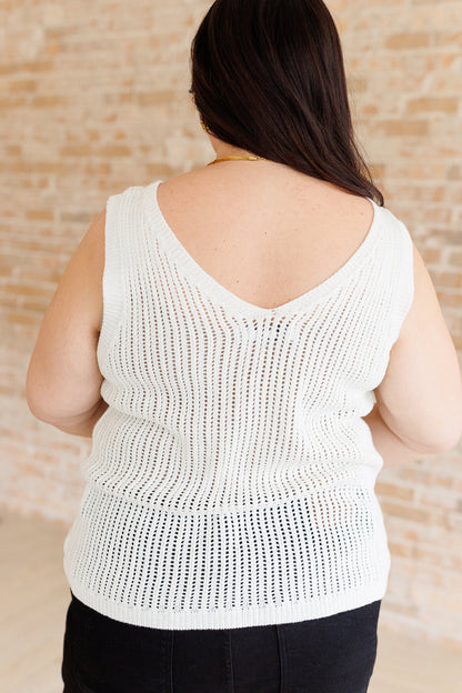 Person wearing a white sleeveless knitted top against a beige brick wall.