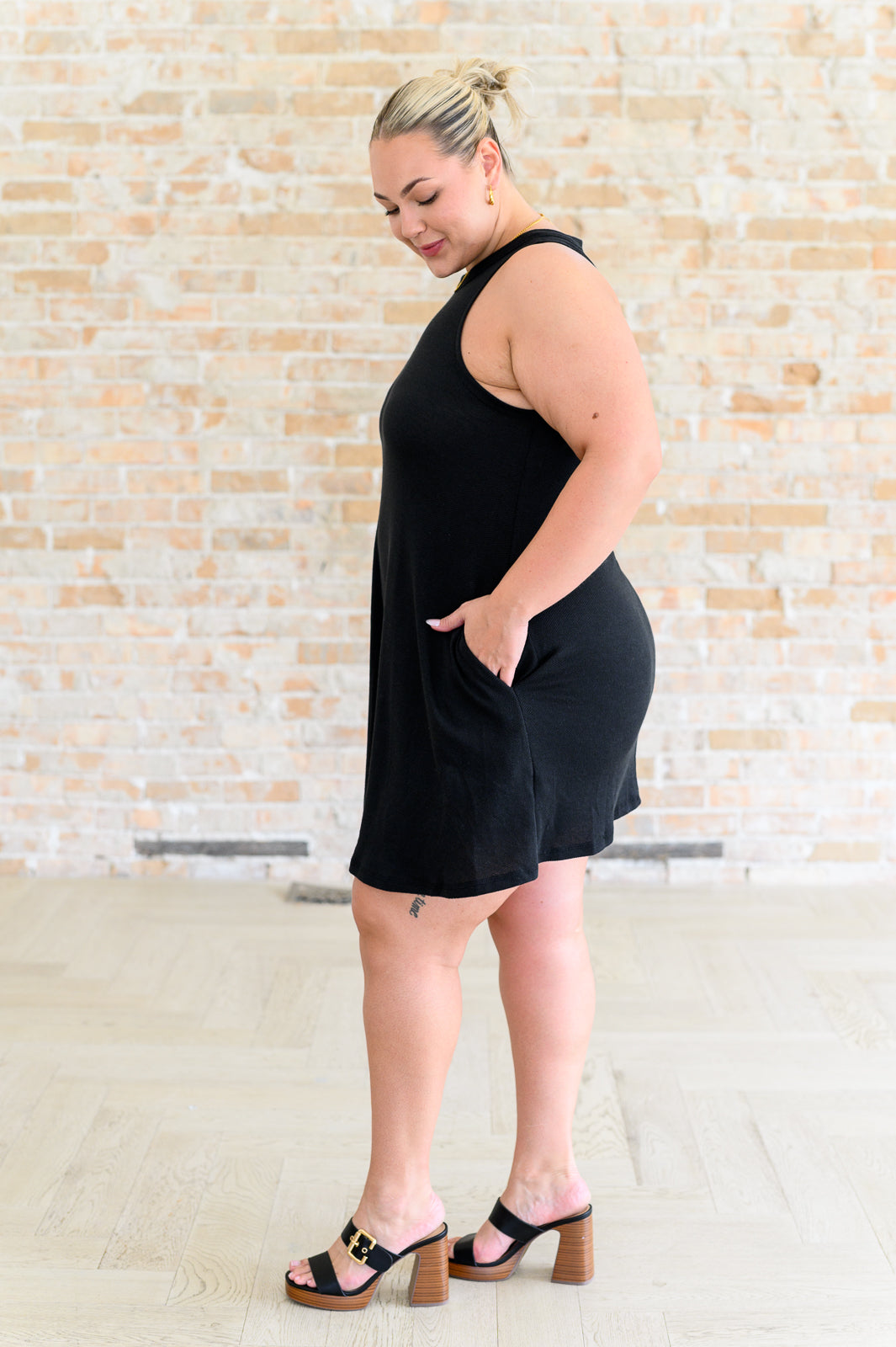 A woman standing in front of a brick wall wearing a black sleeveless knee-length dress with a round neckline and crisscross cutout at the back.
