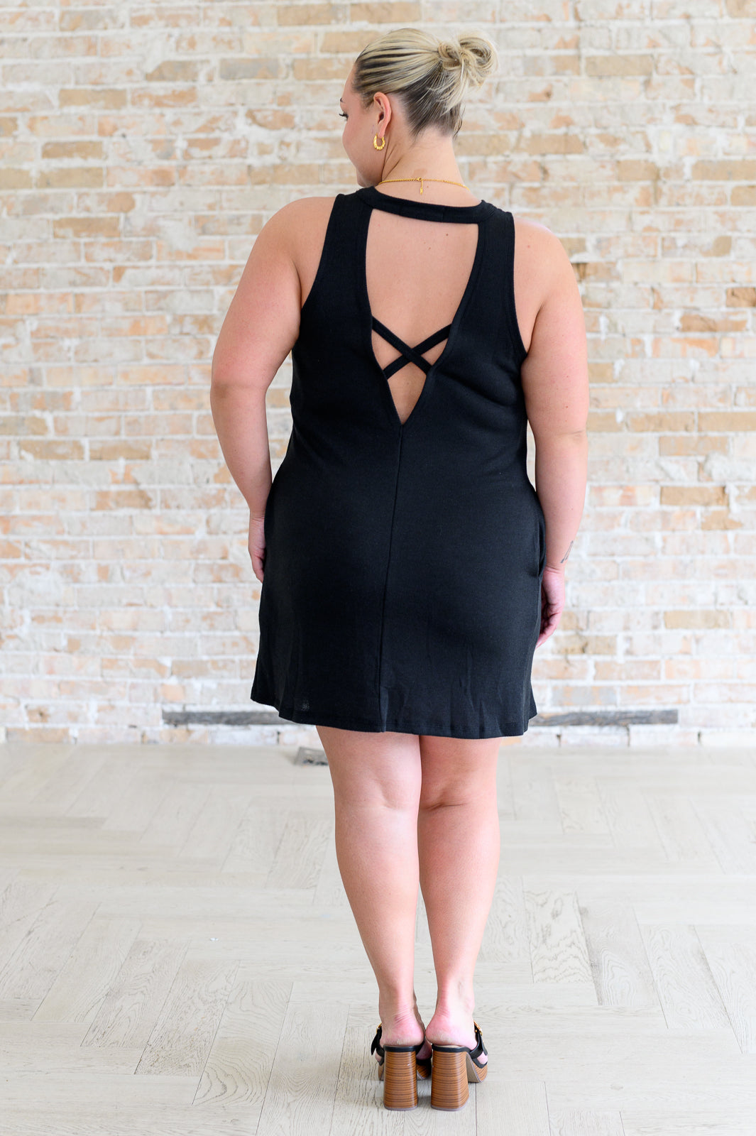 Woman wearing a black dress with criss-cross back design against a light brick wall.