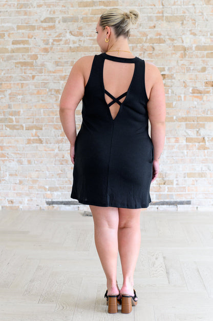 Woman wearing a black dress with criss-cross back design against a light brick wall.