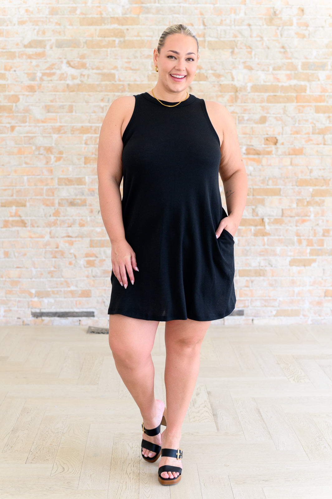 Woman wearing a black sleeveless dress against a light brick wall.