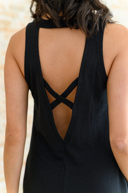 A woman standing in front of a brick wall wearing a black sleeveless knee-length dress with a round neckline and crisscross cutout at the back.