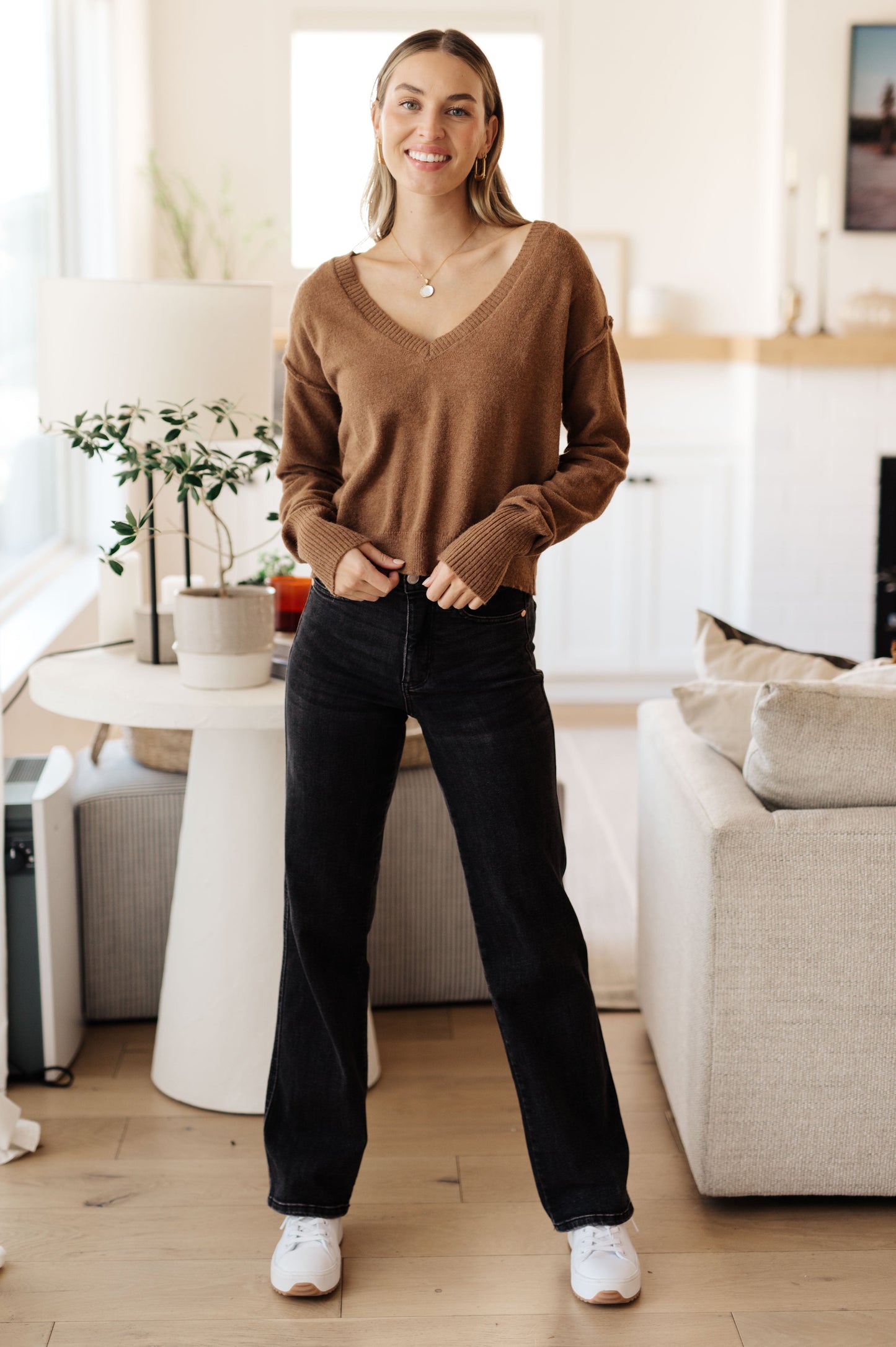 Woman standing in a modern living room wearing a brown sweater and black pants.