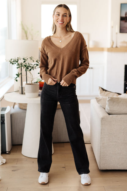 Woman standing in a modern living room wearing a brown sweater and black pants.